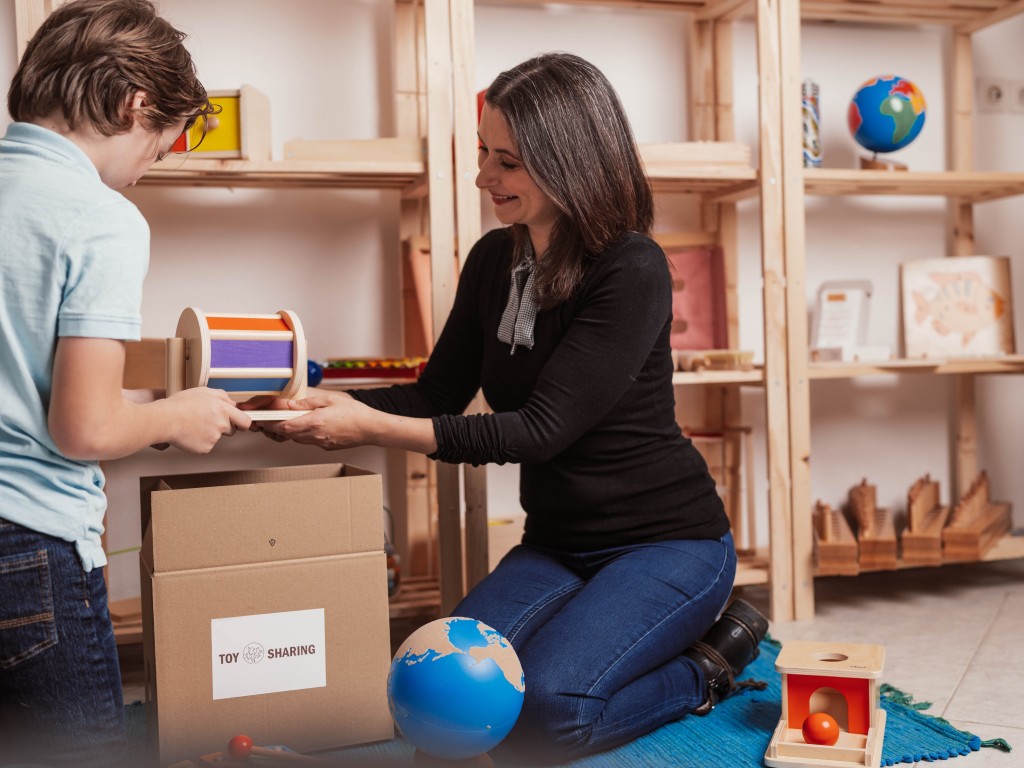 Woman giving a wooden Montessori toy to a child next to a toy rental box.
